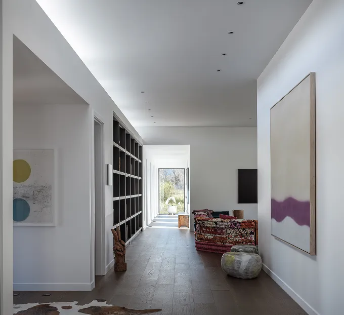 Modern hallway with white walls, wooden floor, built-in dark shelving, colorful patterned sofa, and abstract artwork on walls leading to a bright glass door.