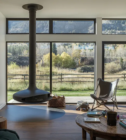 Modern living room with a suspended black fireplace, butterfly chair, wooden coffee table with books, and large windows showing a rural landscape with trees and mountains.