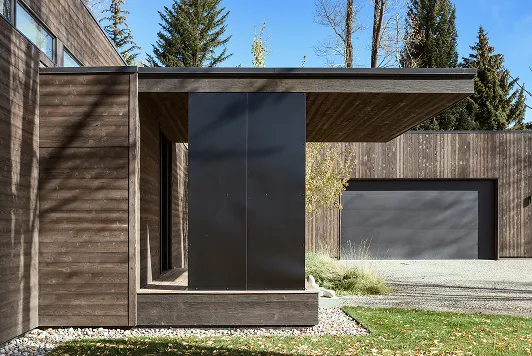 Modern house exterior with horizontal wooden paneling, a black vertical accent, and a large gray garage door surrounded by trees.