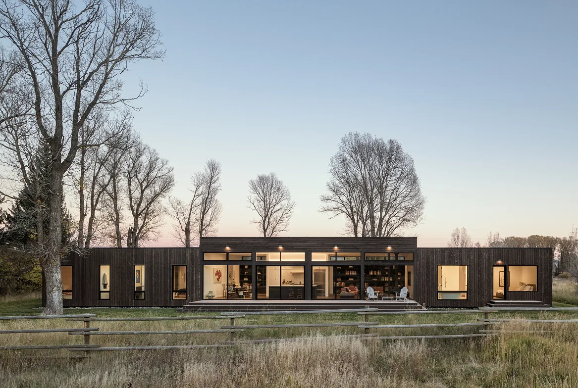 Modern single-story house with large windows and dark wood exterior set against leafless trees and clear sky at dusk.