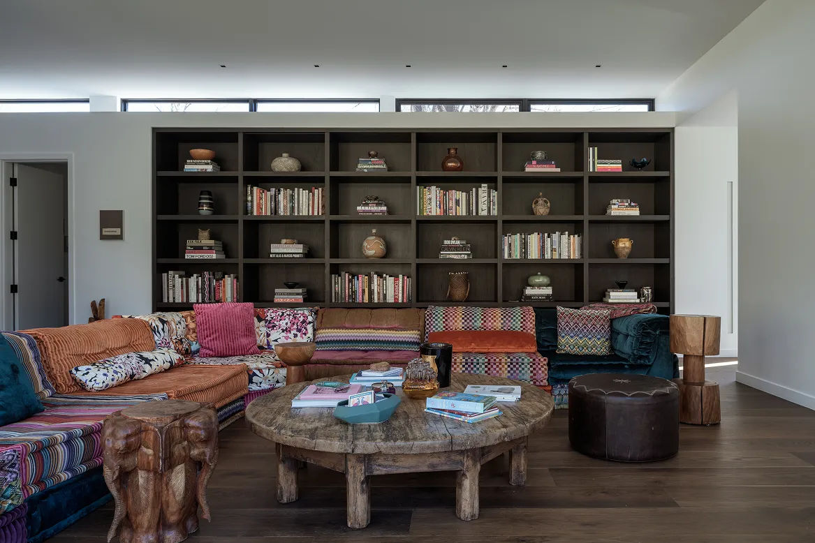 Colorful patchwork sectional sofa with wooden elephant stool and round rustic coffee table in front of dark built-in bookshelves.