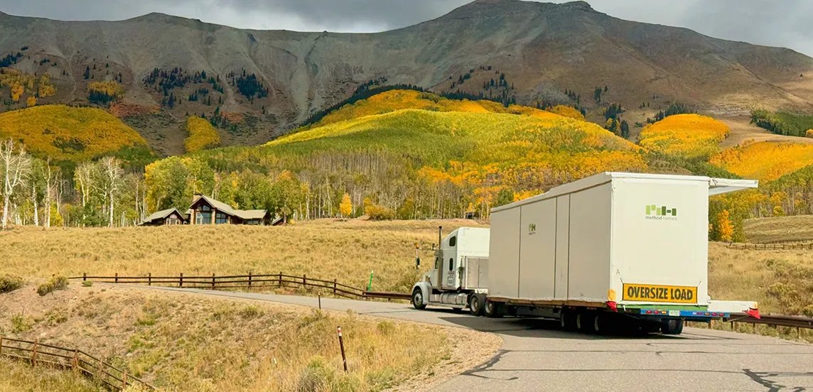 Truck carrying a large white modular home with 'oversize load' sign driving on a winding road surrounded by dry grass and colorful autumn trees against mountain backdrop.