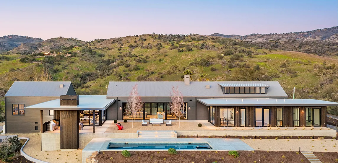 Modern single-story house with large windows, outdoor seating area, and a rectangular swimming pool, set against green rolling hills at sunset.