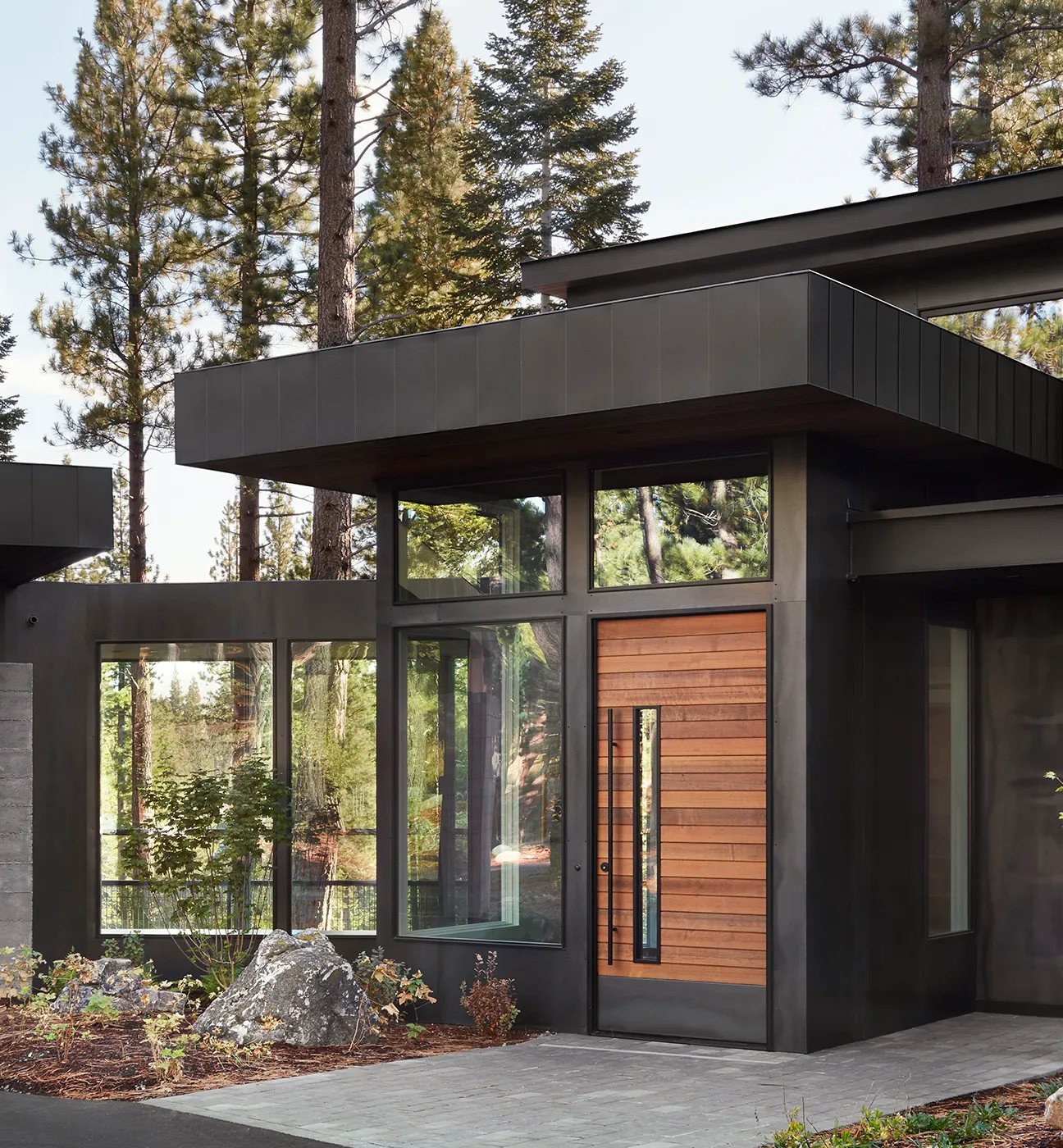 Modern entryway with large wooden front door and tall glass windows reflecting surrounding pine trees.