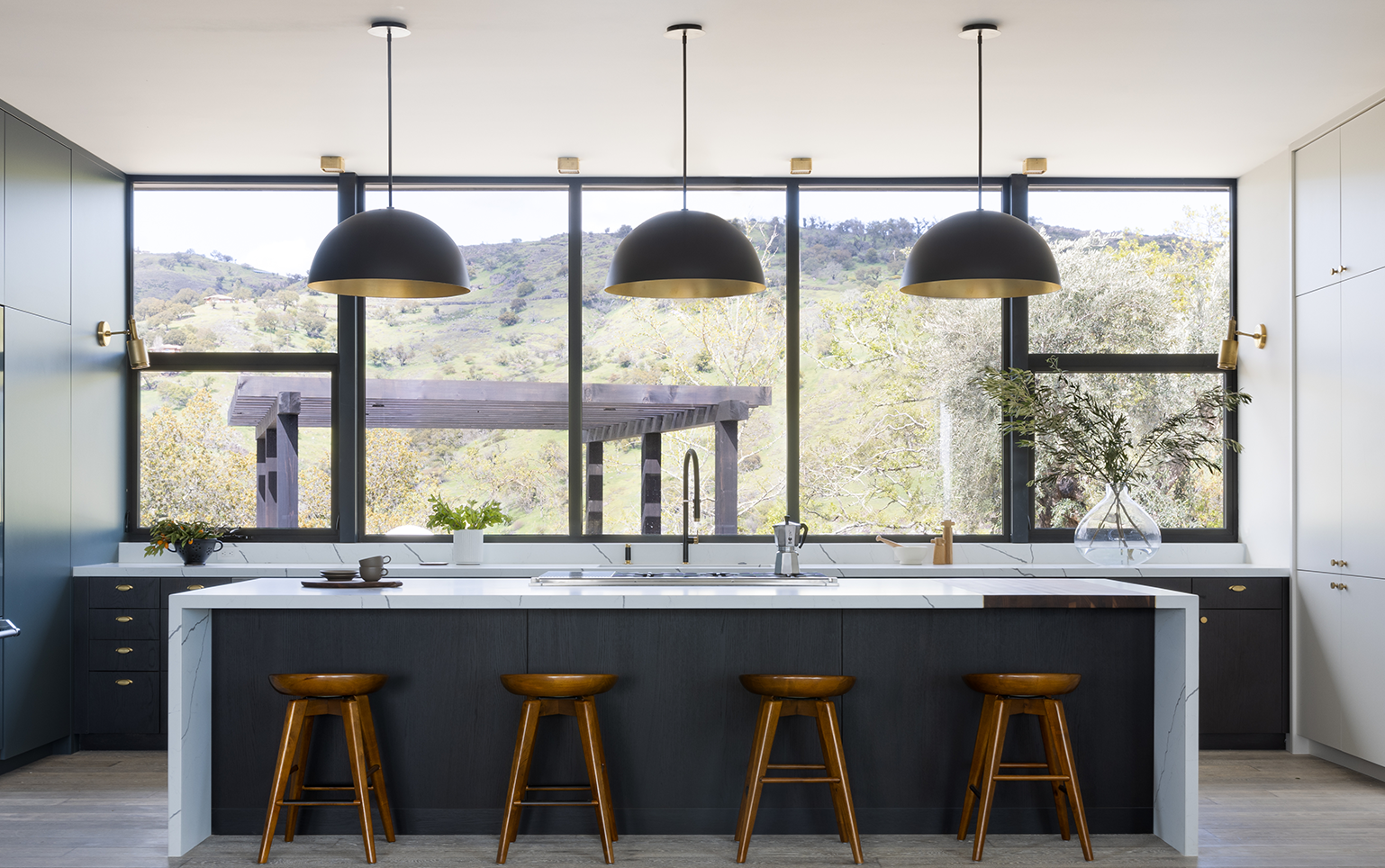 Modern kitchen with large island, four wooden stools, three black pendant lights, and a view of green hills through wide windows.