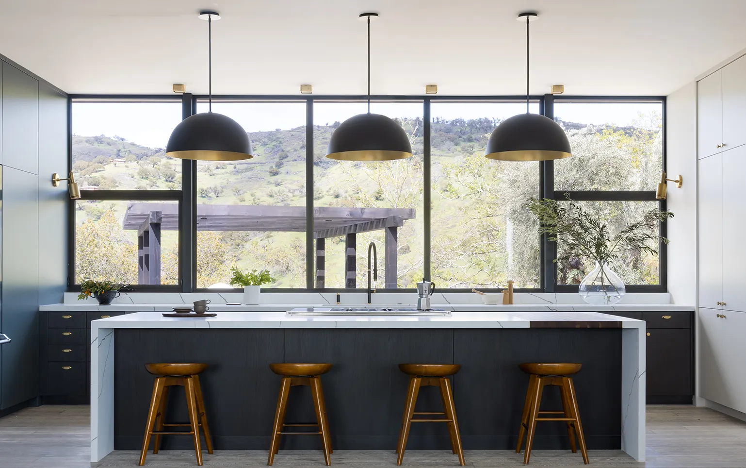 Modern kitchen with large island, four wooden stools, three black pendant lights, and a view of green hills through wide windows.