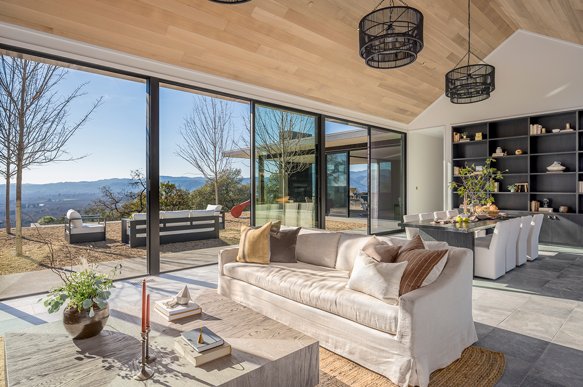 Bright open living and dining area with beige sofa, wooden coffee table, large sliding glass doors overlooking outdoor seating and scenic mountain views.