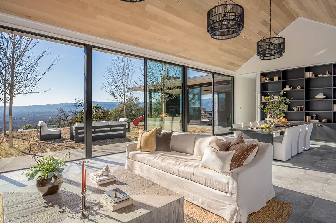 Bright open living and dining area with beige sofa, wooden coffee table, large sliding glass doors overlooking outdoor seating and scenic mountain views.
