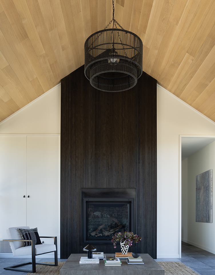 Modern living room with vaulted light wood ceiling, dark wood-paneled fireplace, concrete coffee table with books and flower vase, and a gray armchair.