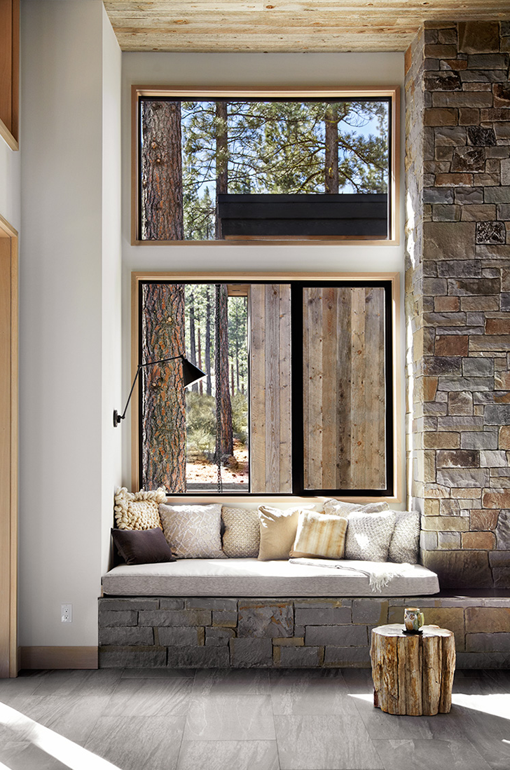 Cozy reading nook with cushioned bench, assorted pillows, stone wall, wooden ceiling, and large windows showing a forest view.