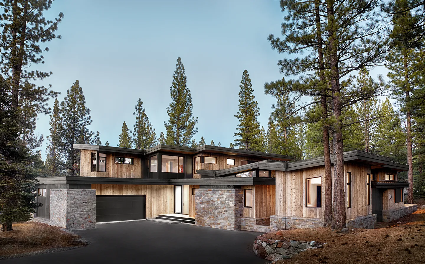 Modern two-story house with wooden and stone exterior, illuminated warm interior lights, surrounded by pine trees at dusk.