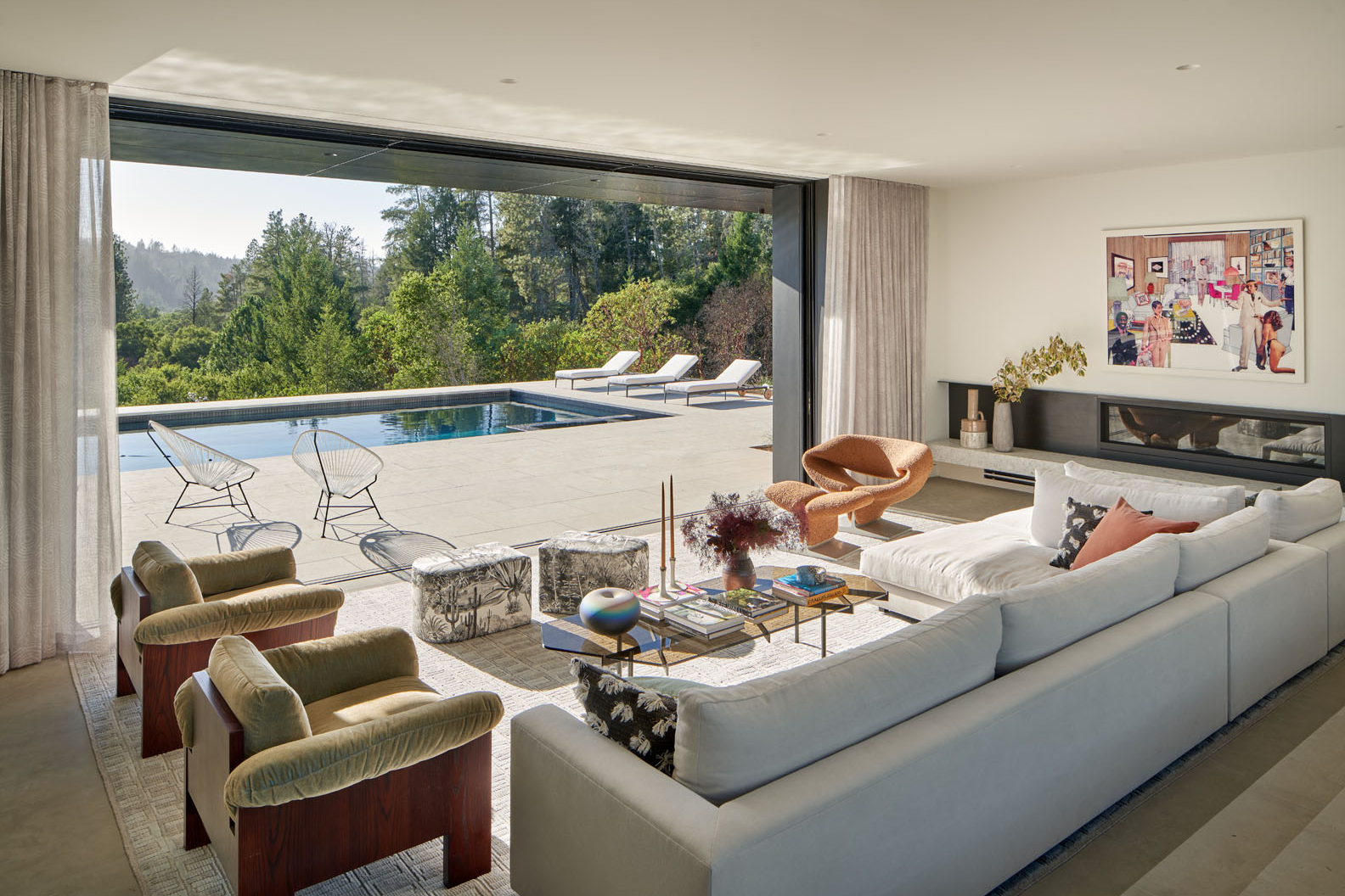 Modern living room with large white sectional sofa, velvet armchairs, and a glass coffee table, opening to a patio with pool and lounge chairs surrounded by trees.