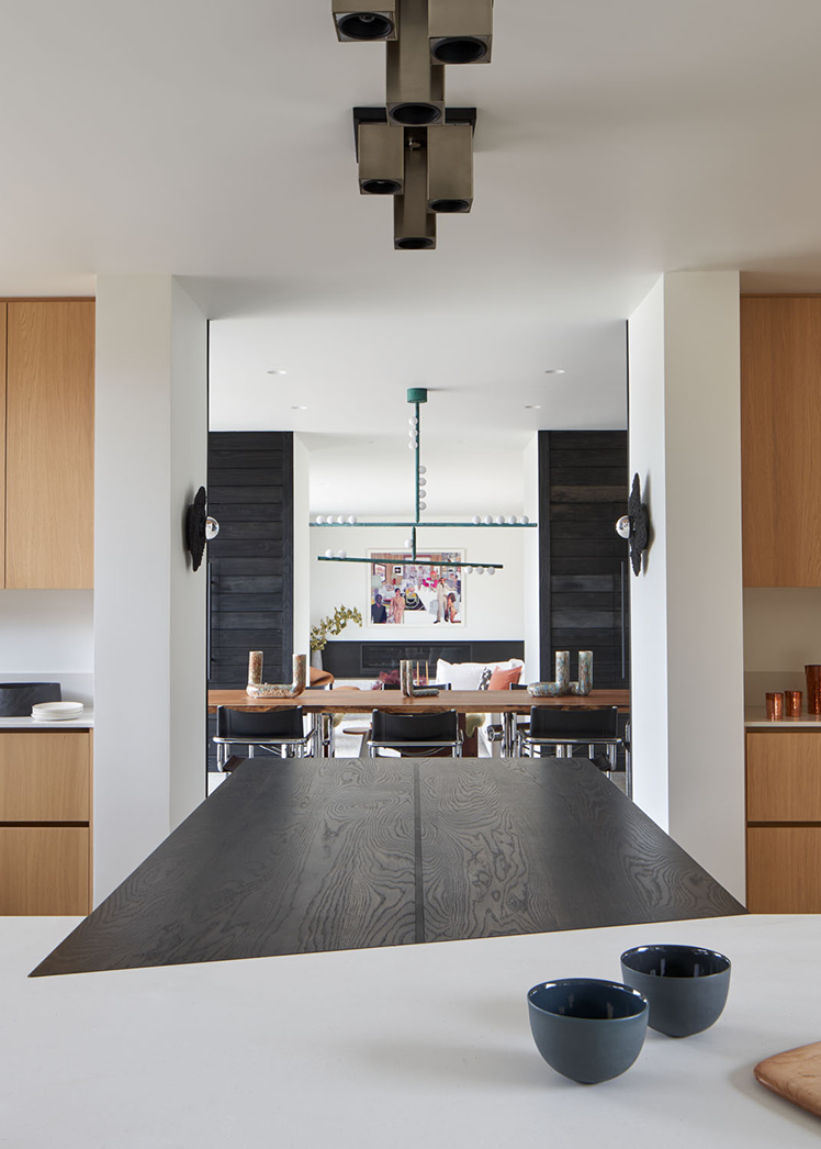 Modern open kitchen with light wood cabinets, a dark wooden dining table, and contemporary lighting fixtures, looking into a seating area with black paneled walls and artwork.