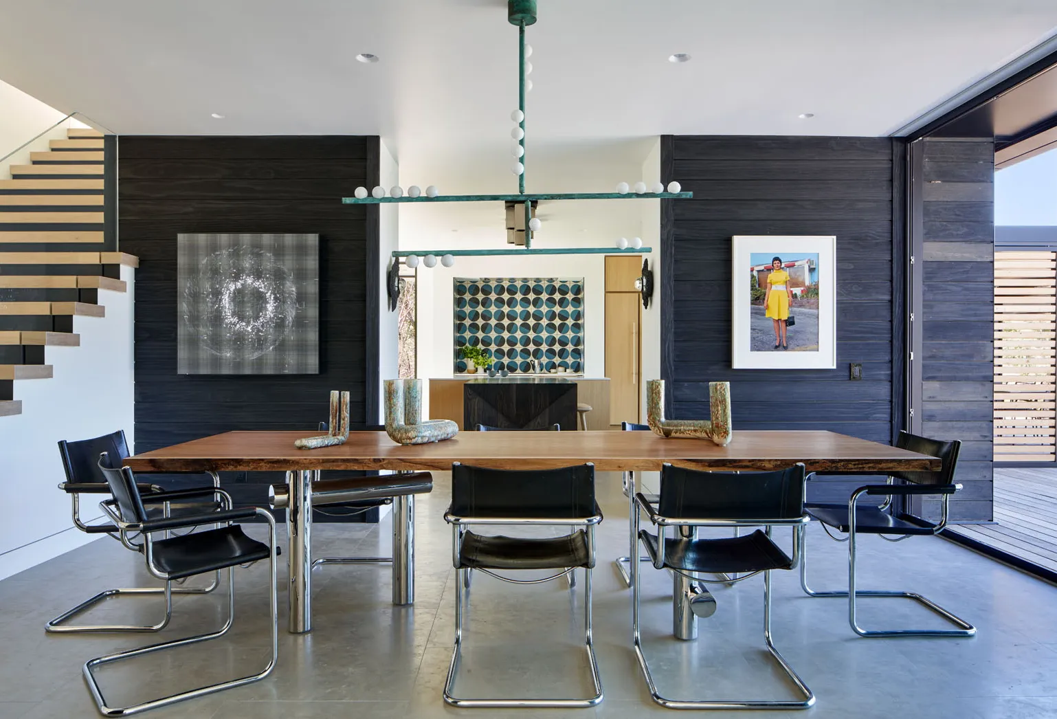 Modern dining room with a wooden table, six black leather chairs with metal frames, dark wood paneled walls, abstract and portrait artwork, and an open view to a kitchen and outdoor deck.