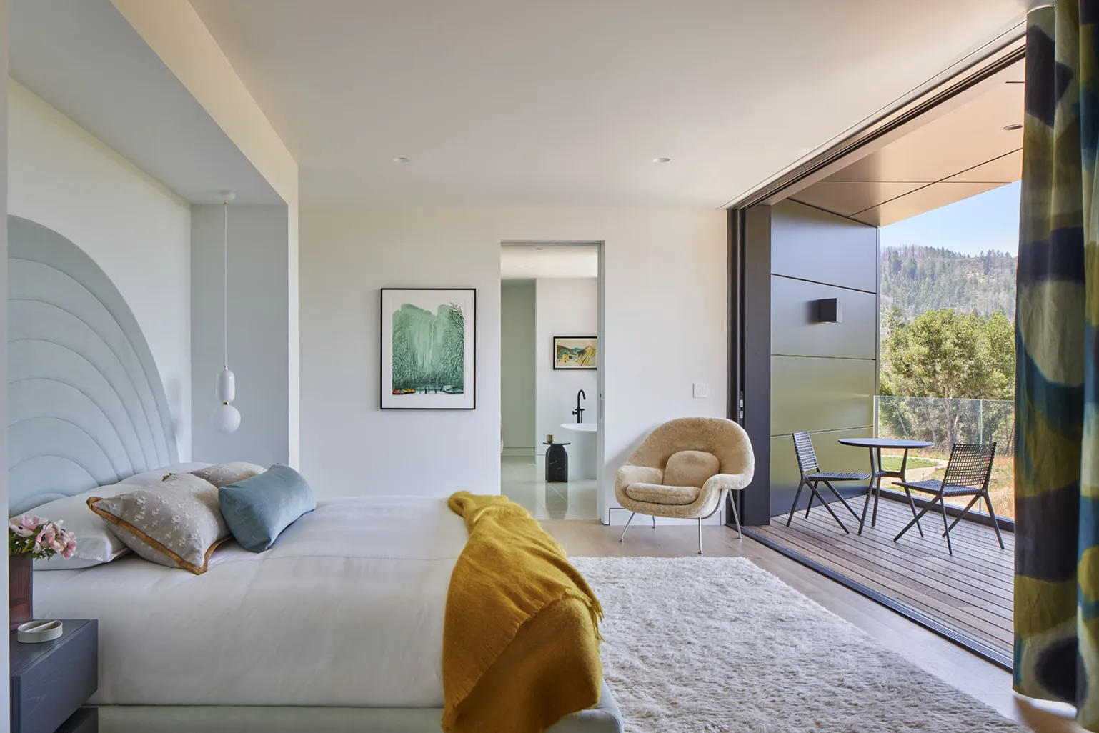 Modern bedroom with a large bed featuring a blue curved headboard, a mustard yellow throw blanket, a beige plush chair, and a balcony with a table and two chairs overlooking trees and hills.