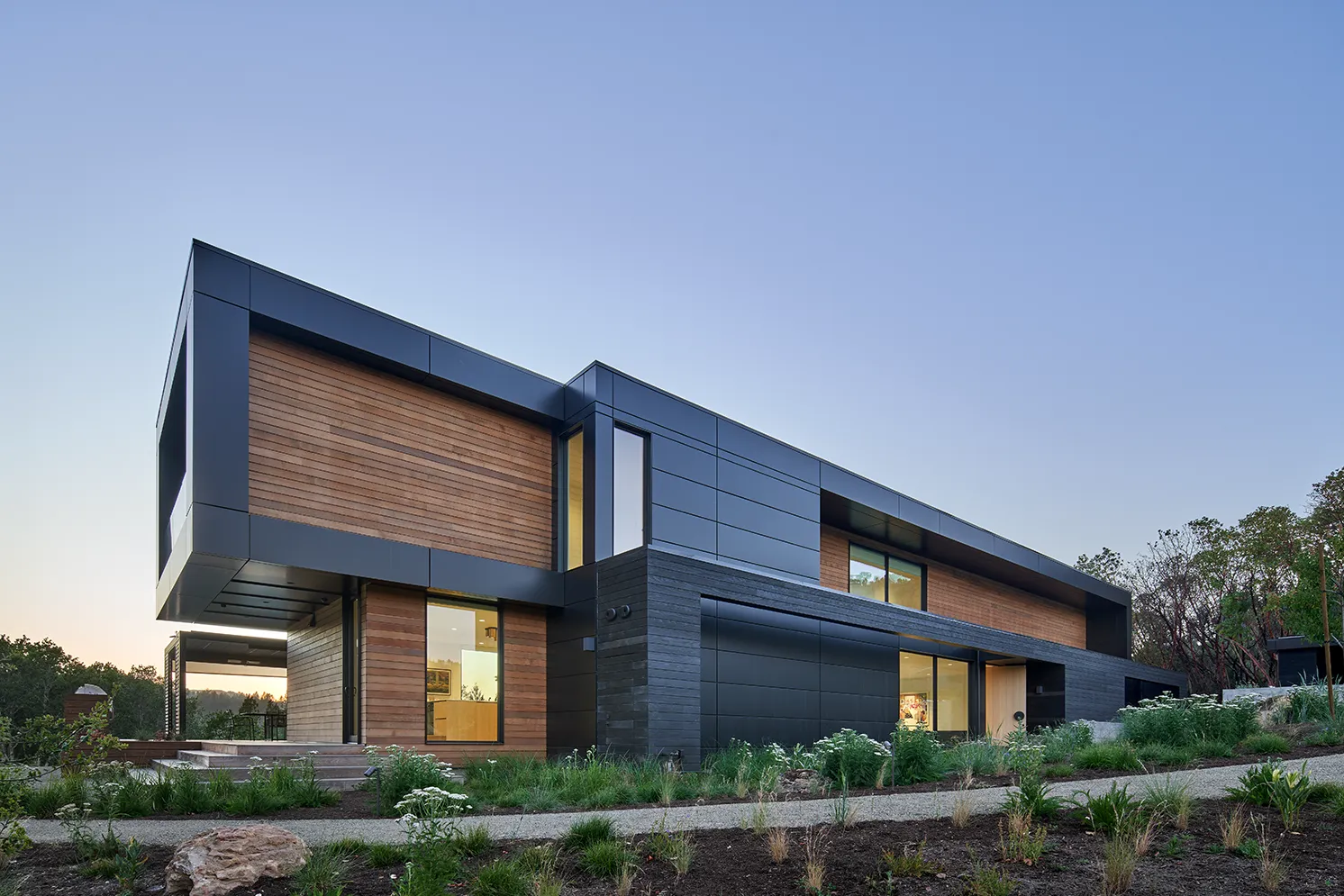 Modern two-story house with black and wooden panel exterior, large windows, and landscaped garden at twilight.