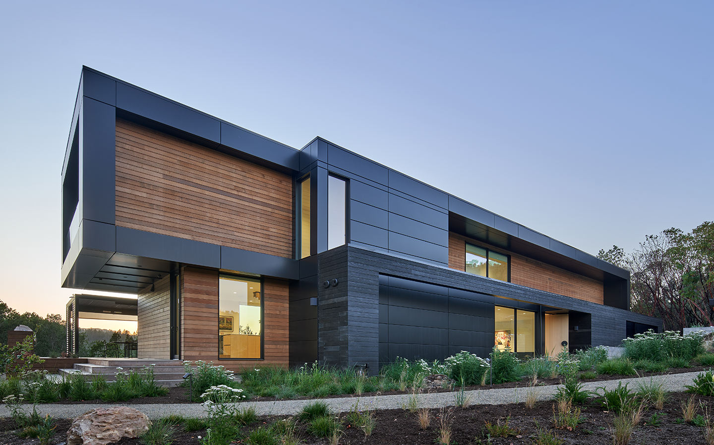Modern two-story house with black and wooden panel exterior, large windows, and landscaped garden at twilight.