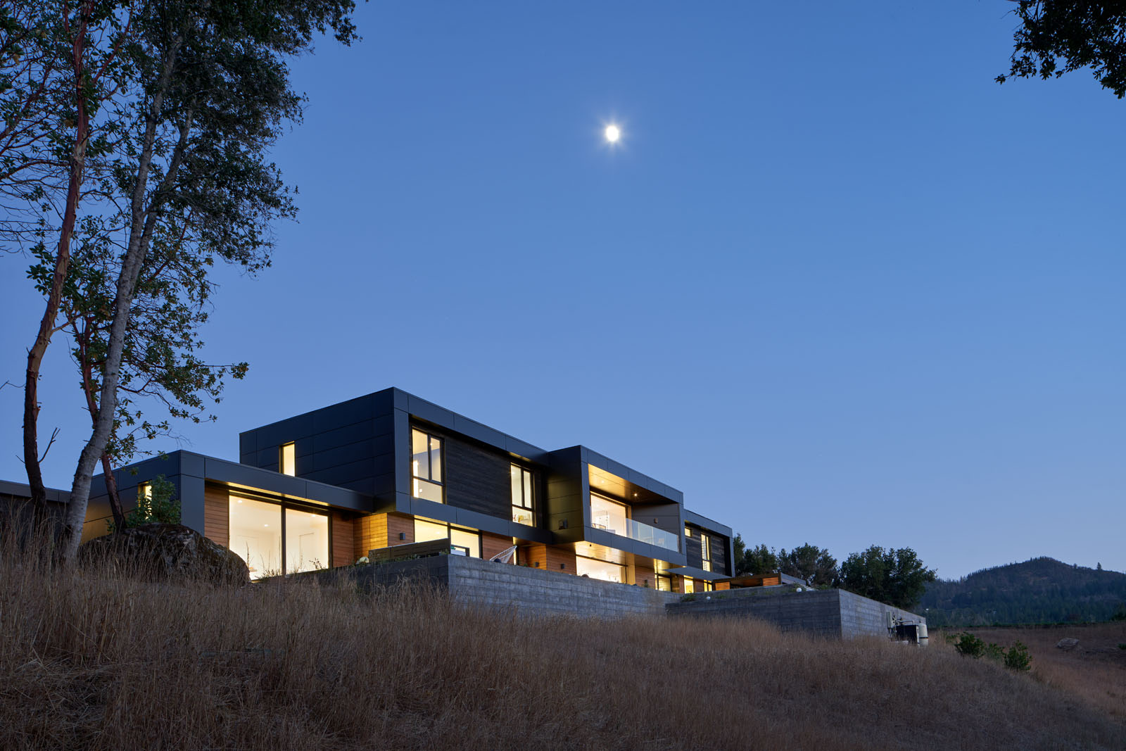 Modern two-story house with large windows illuminated at dusk under a clear sky with the moon visible.
