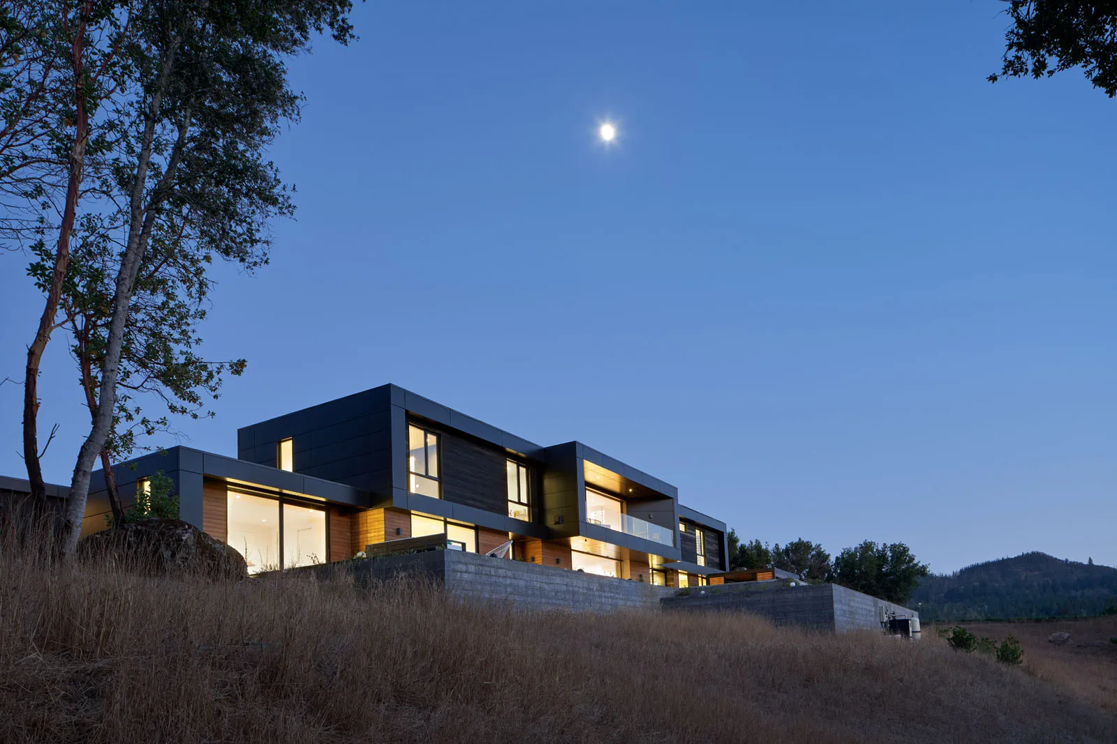 Modern two-story house with large windows illuminated at dusk under a clear sky with the moon visible.