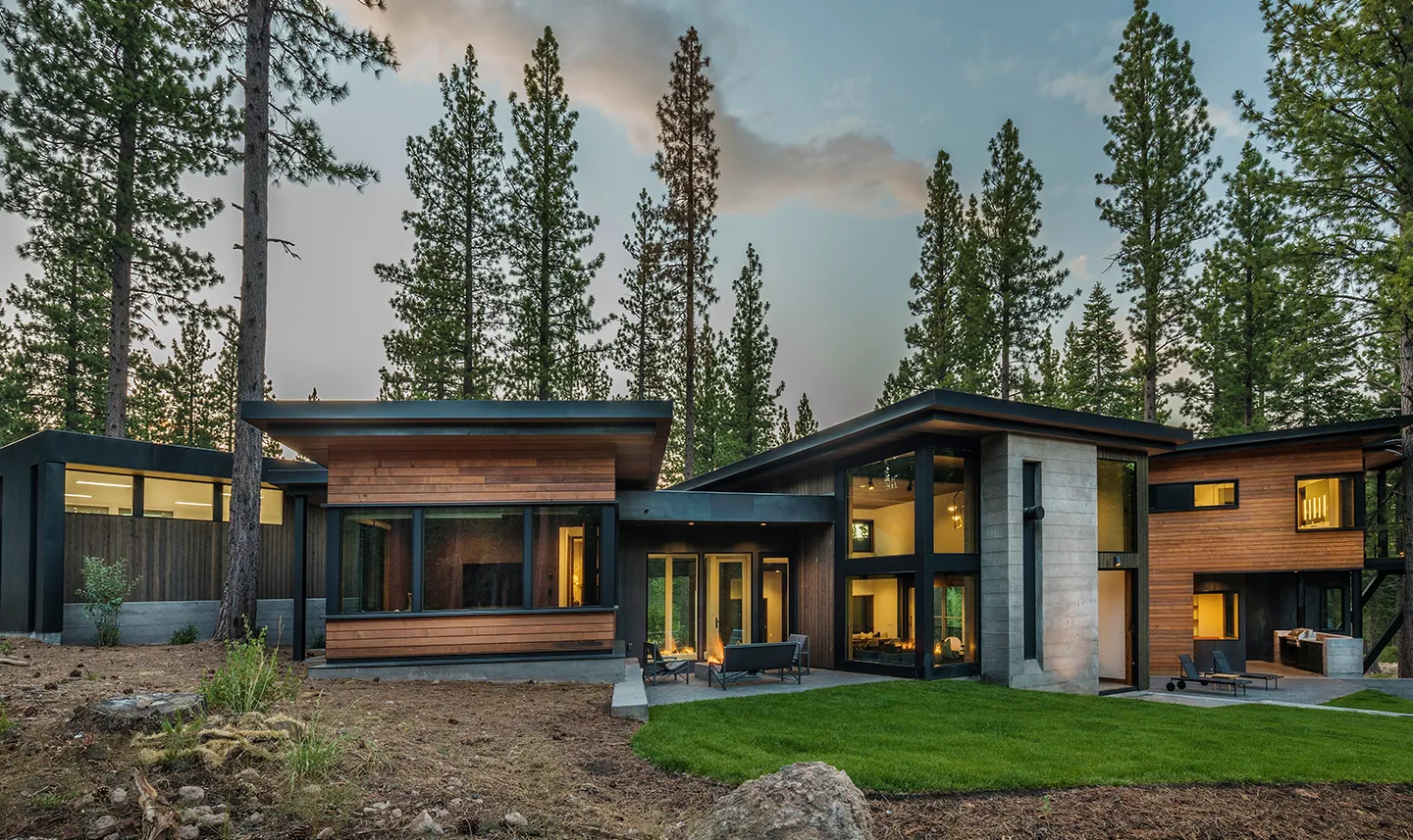 Modern single-story house with large windows, wood and concrete exterior, outdoor seating, and surrounded by tall pine trees at dusk.