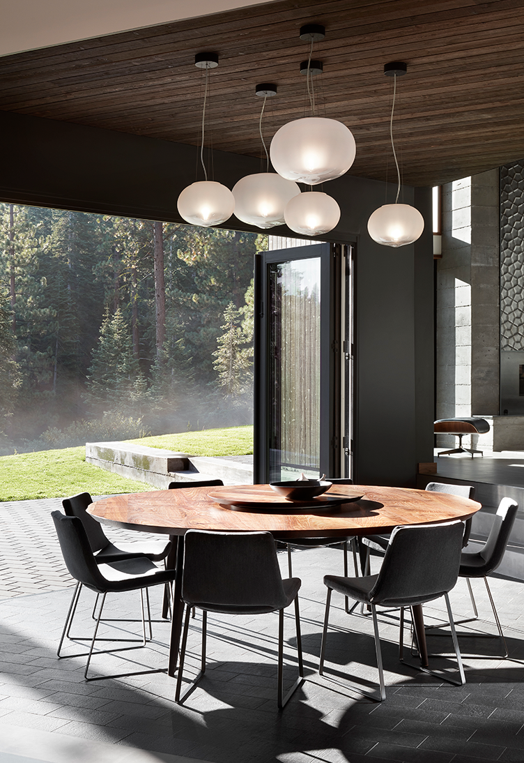 Modern dining area with a round wooden table surrounded by six gray chairs, large glass doors opening to an outdoor green forest view, and hanging white globe pendant lights.