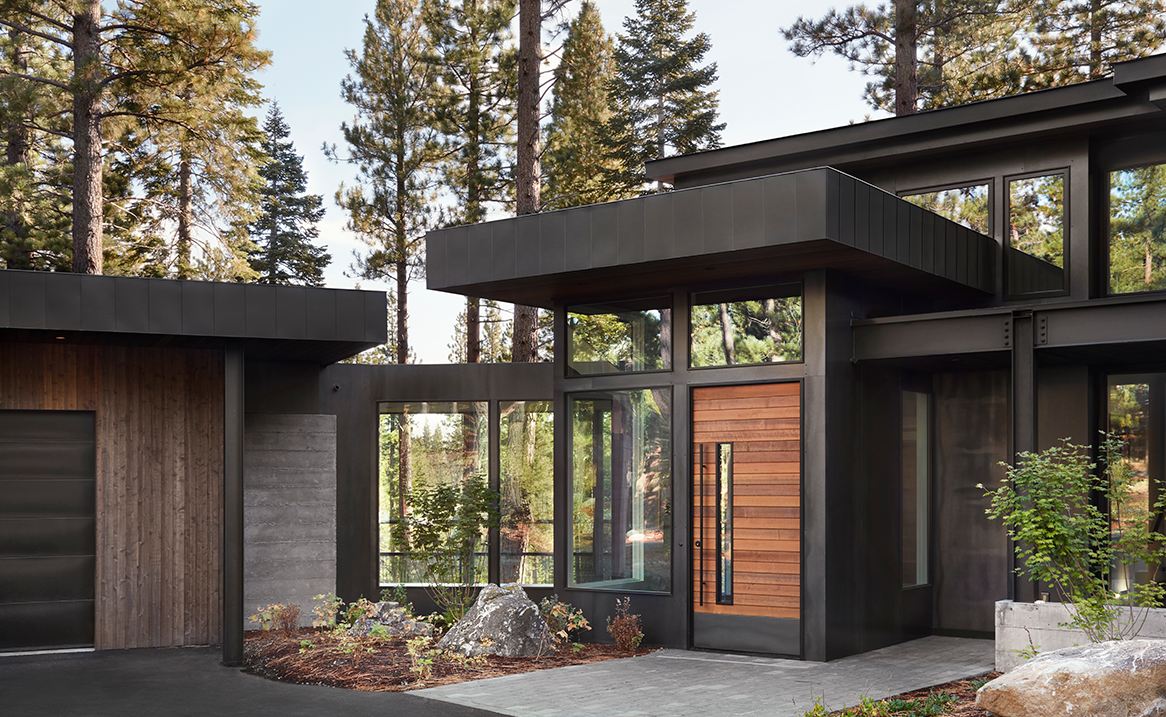 Modern house entrance with large wooden front door, black metal framing, floor-to-ceiling windows, and surrounding tall pine trees.