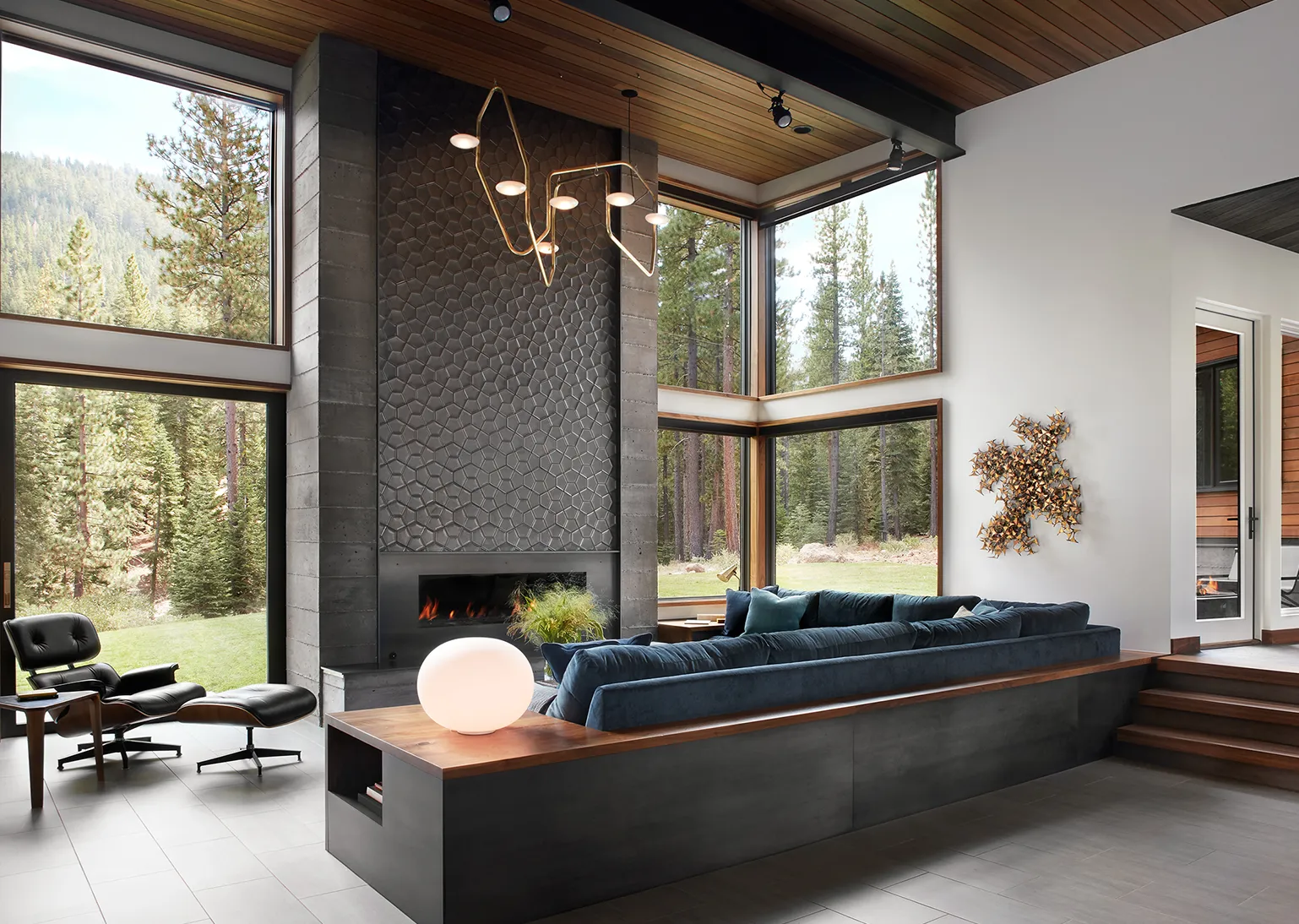 Modern living room with large corner windows showing forest views, a dark blue sectional sofa, black lounge chair with ottoman, and a fireplace with textured dark wall behind it.