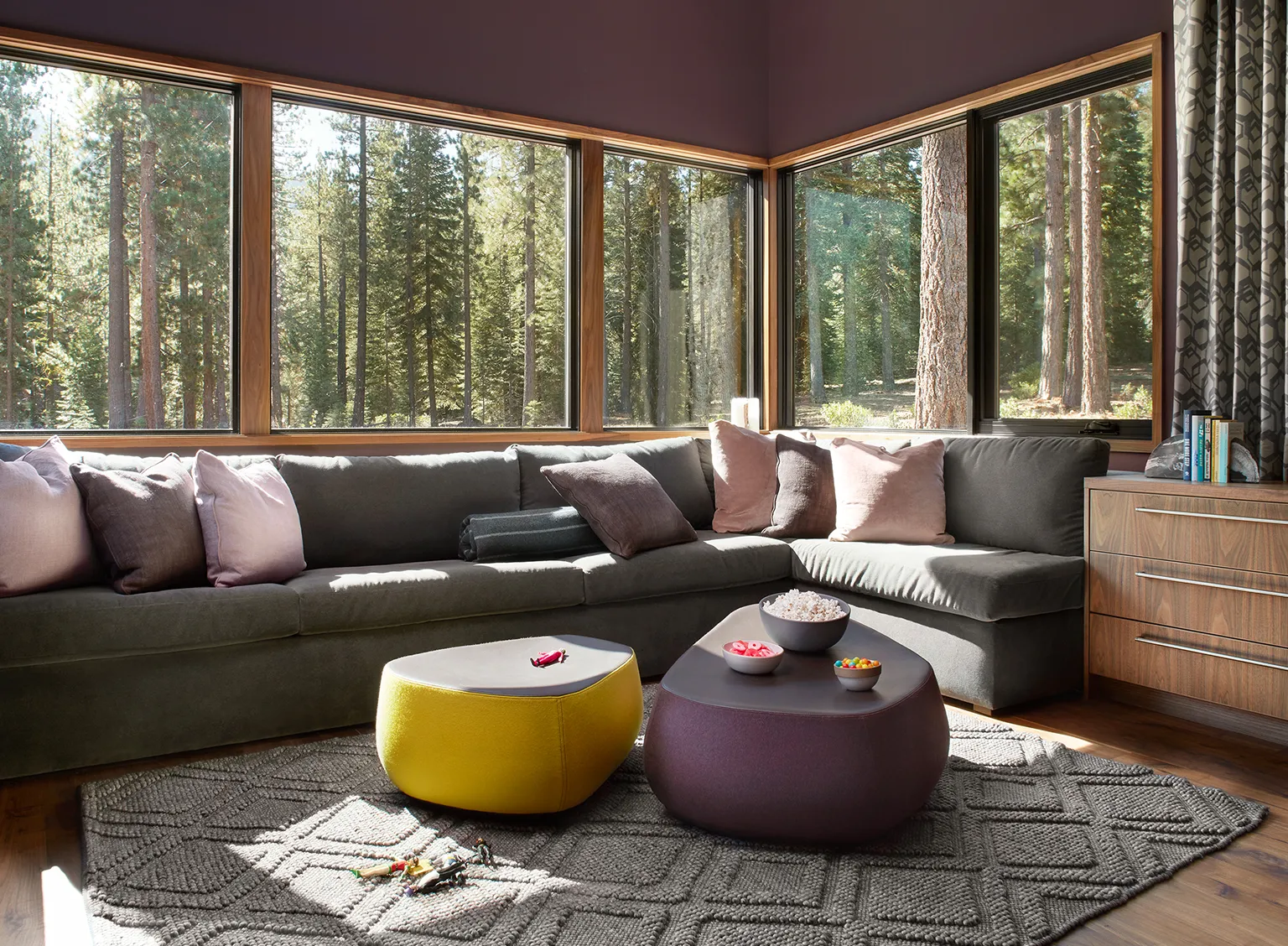 Cozy living room corner with large gray sectional sofa, pink and gray pillows, two modern ottomans with snacks, children’s toys on a textured rug, and large windows showing a forest view.