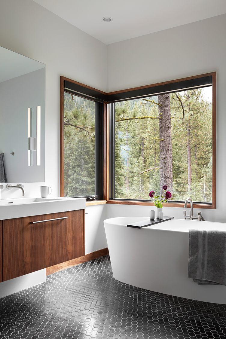 Modern bathroom with a white freestanding bathtub, black hexagonal tile floor, wood-paneled vanity, large corner window showing forest view, and a gray towel hanging on the tub.
