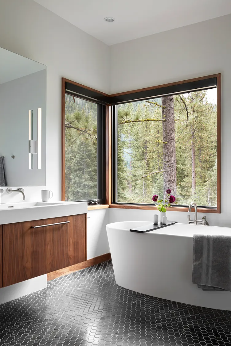 Modern bathroom with a white freestanding bathtub, black hexagonal tile floor, wood-paneled vanity, large corner window showing forest view, and a gray towel hanging on the tub.