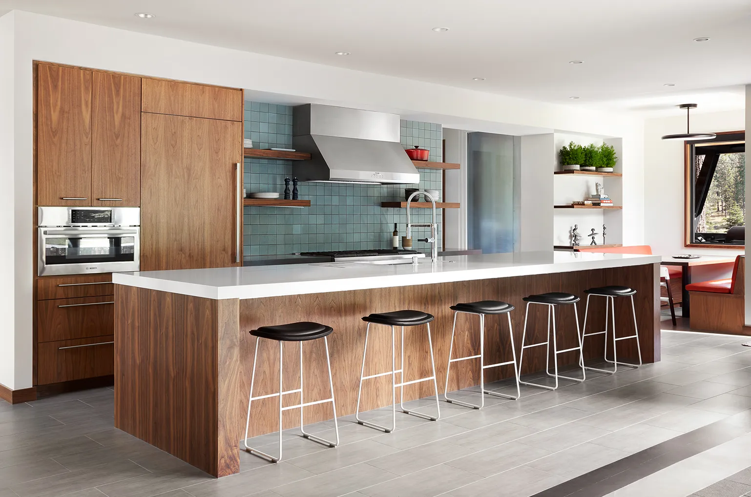 Modern kitchen with a long island featuring a white countertop, five black bar stools, wooden cabinets, stainless steel appliances, and blue square tile backsplash.