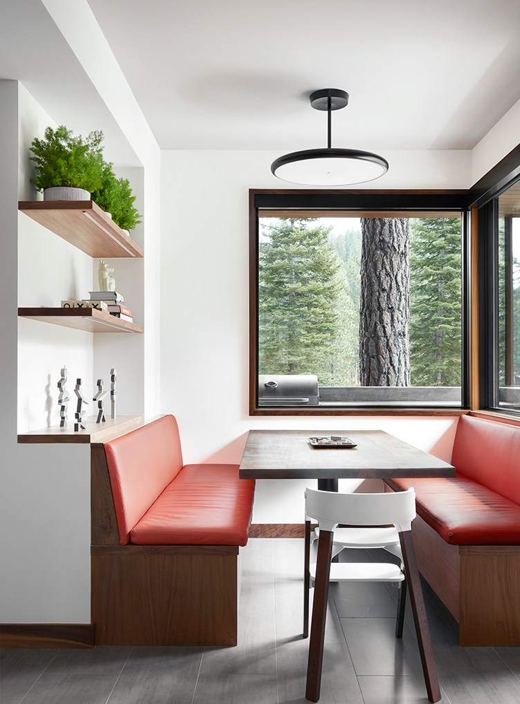 Modern dining nook with red leather booth seating, wooden table, white chair, floating shelves with decor, and large window showing pine trees outside.