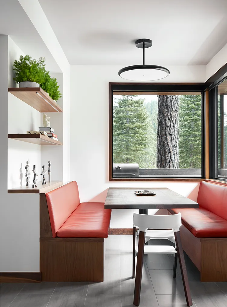 Modern dining nook with red leather booth seating, wooden table, white chair, floating shelves with decor, and large window showing pine trees outside.