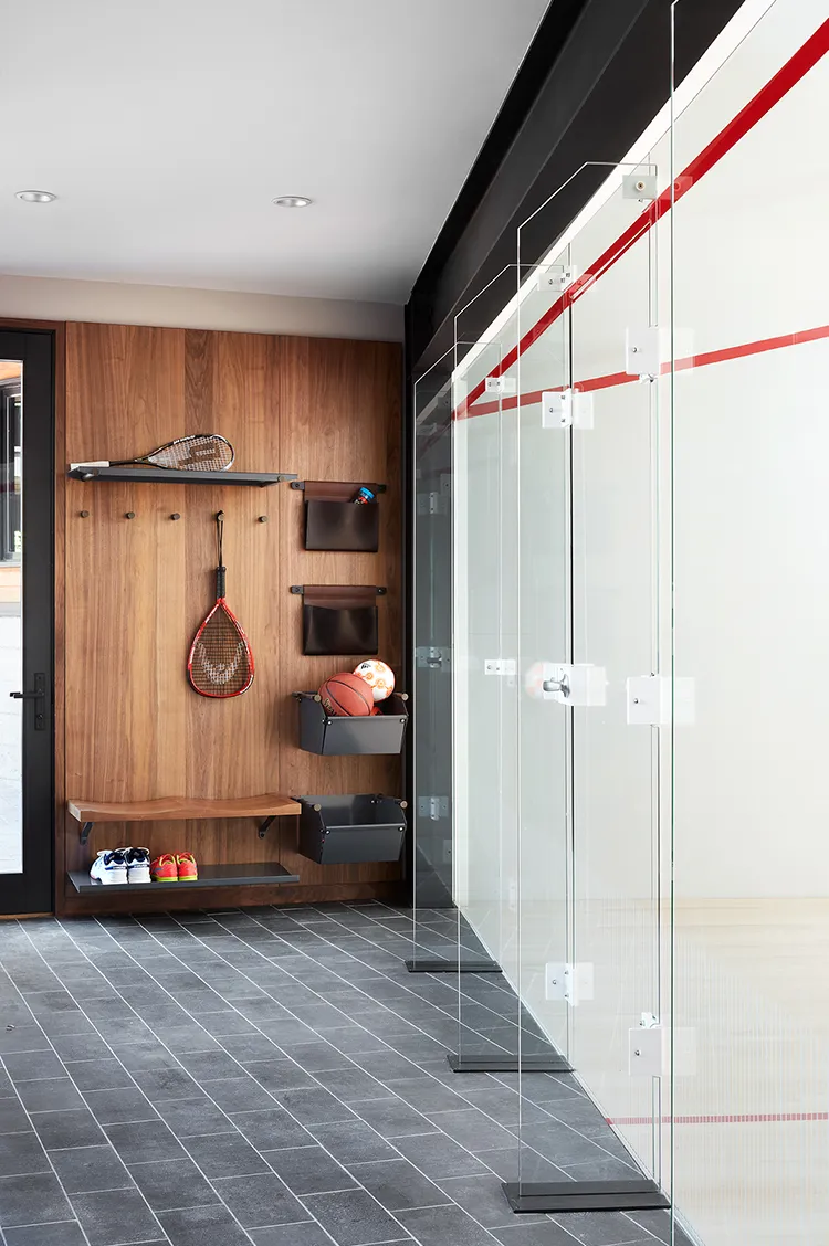 Modern indoor racquetball court with glass walls and a wooden storage area holding rackets, basketball, soccer ball, and shoes.