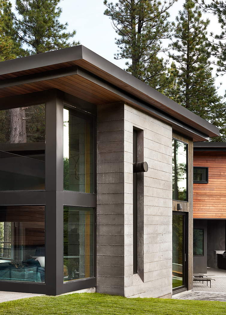 Modern house exterior with large black-framed windows, concrete and wood siding, surrounded by pine trees and green lawn.