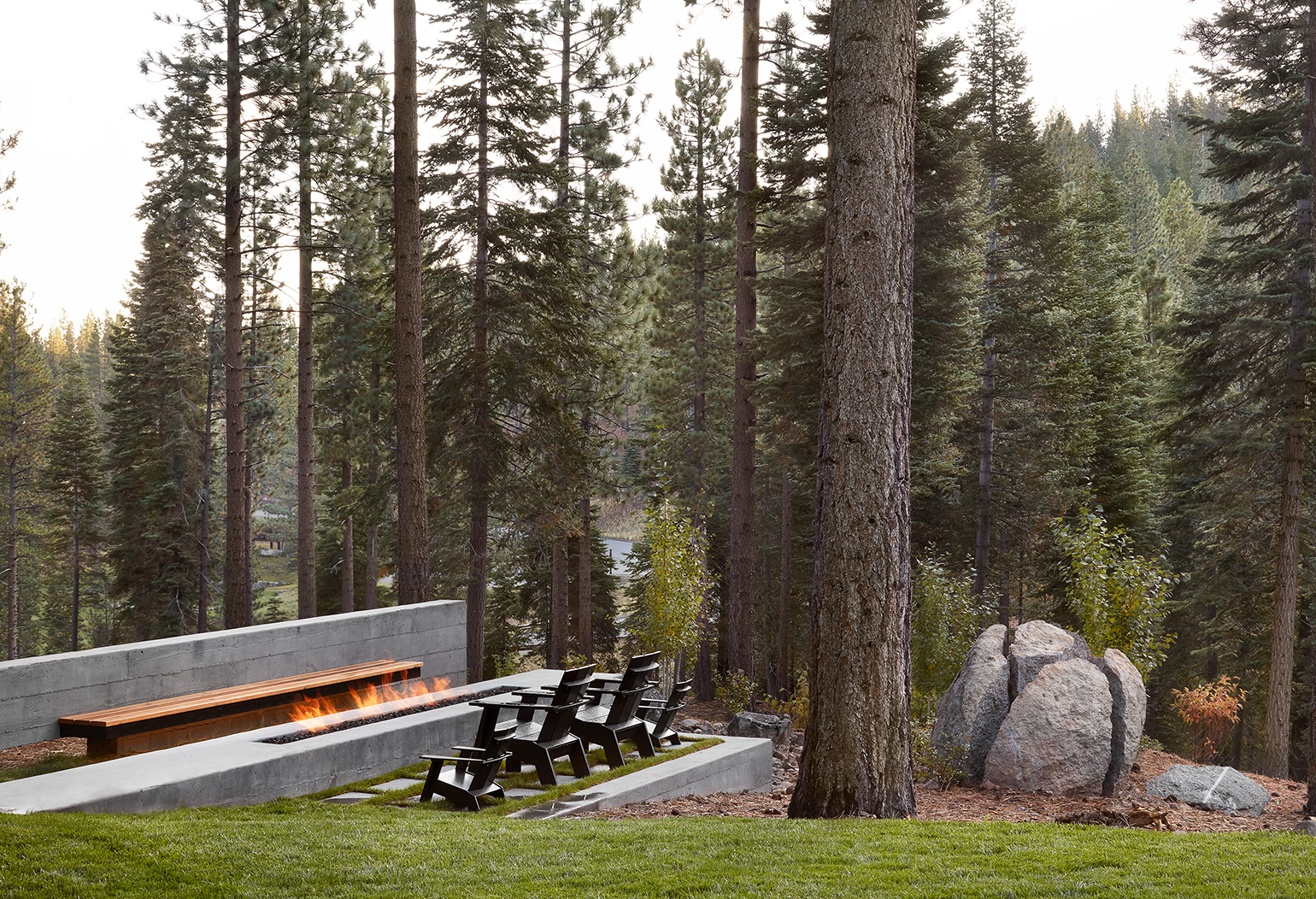 Outdoor seating area with black chairs and a long rectangular fire pit surrounded by tall pine trees.