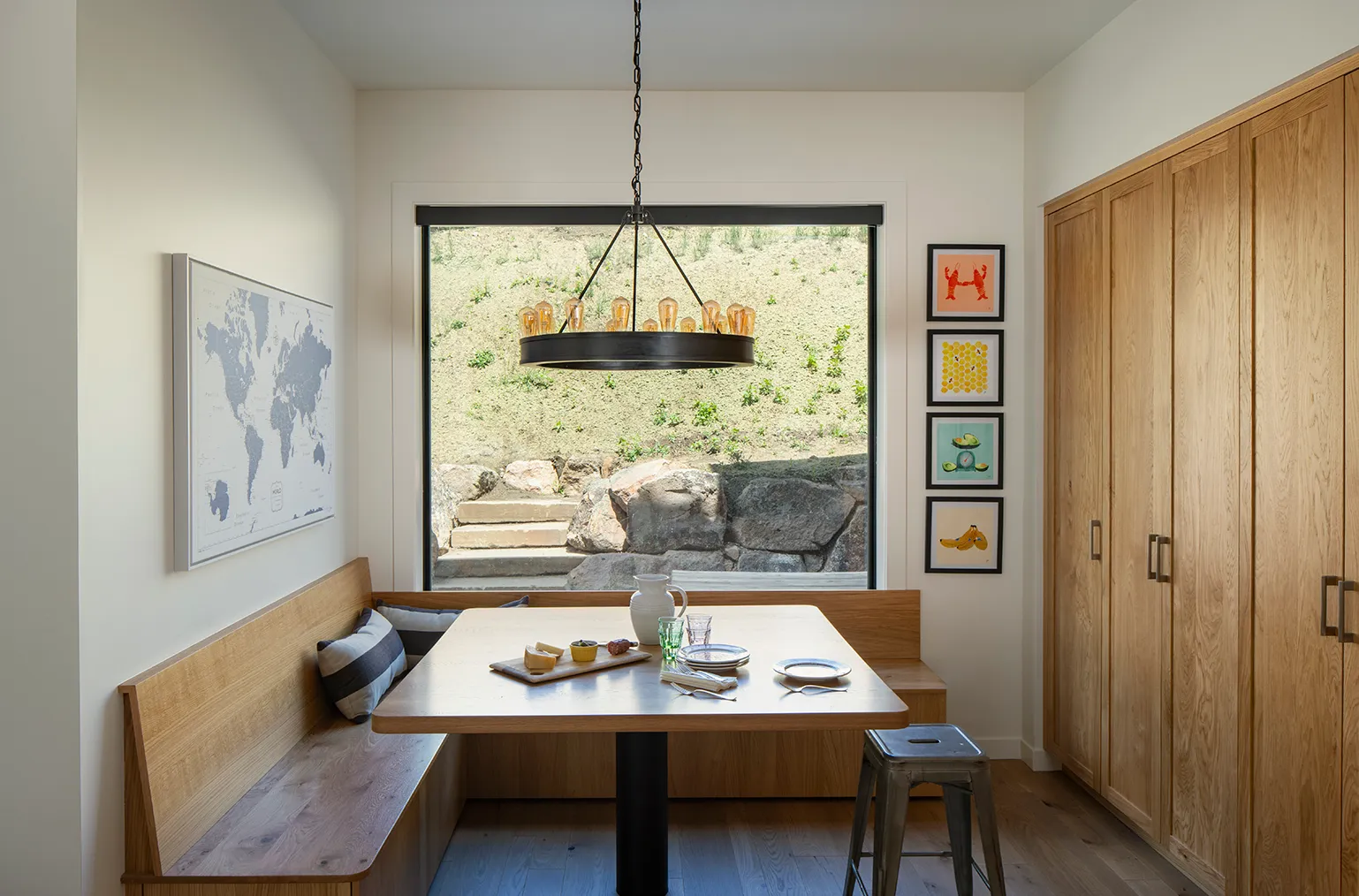 Cozy dining nook with wooden bench seating, a table set with dishes, and a large window overlooking stone steps and greenery.