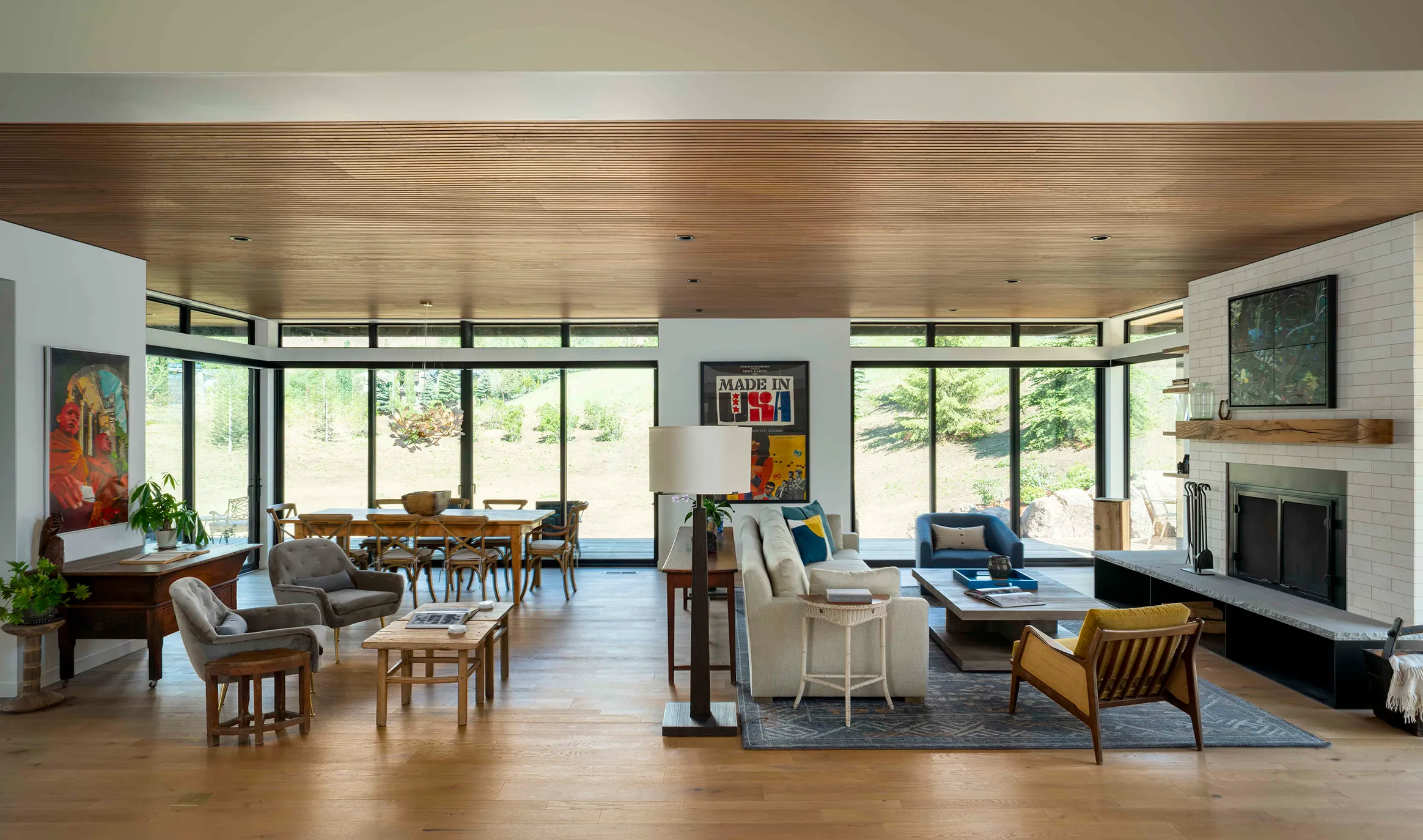 Open living and dining area with wooden floor, modern furniture, large glass windows showing a landscape view, and a fireplace.