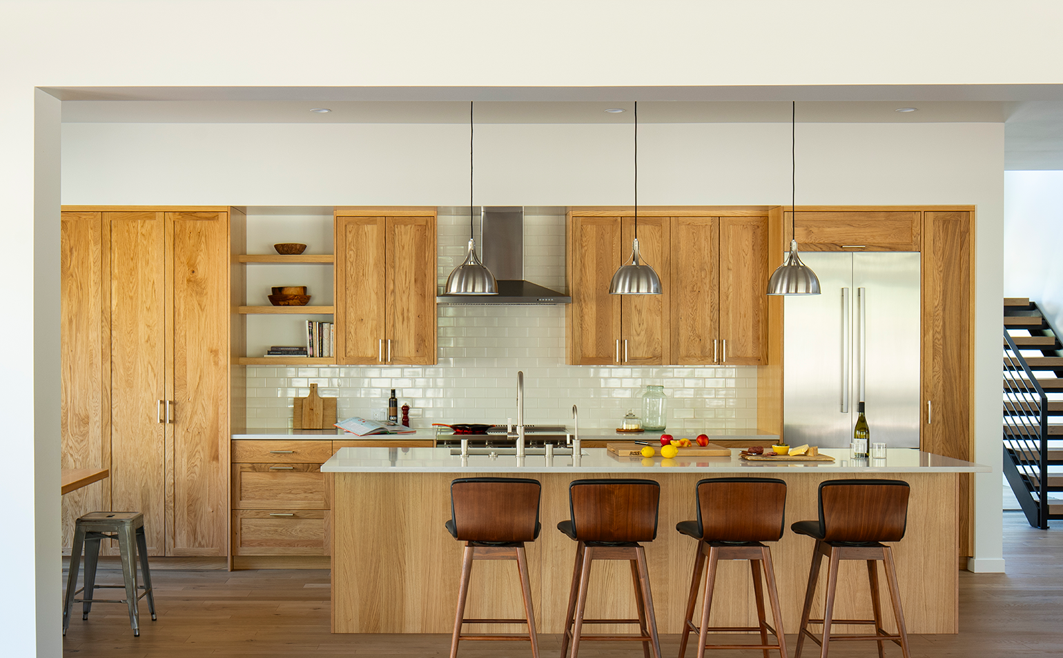 Modern kitchen with wooden cabinets, white countertop island with four wooden stools, stainless steel appliances, and hanging metal pendant lights.
