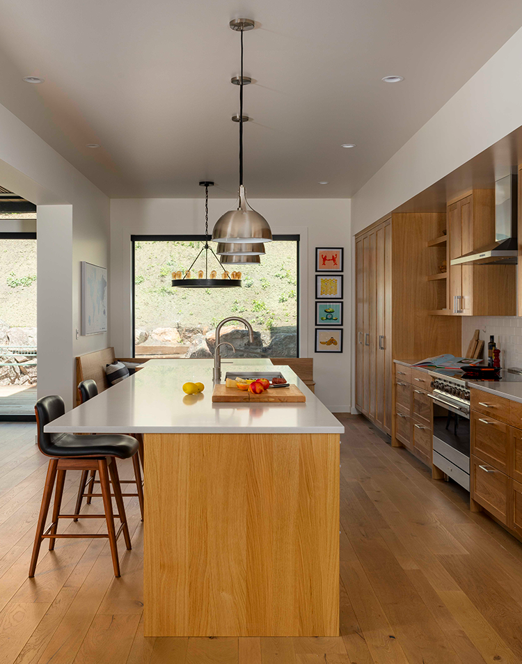 Modern kitchen with wooden cabinets, a large island with white countertop, two black bar stools, hanging pendant lights, and a window showing an outdoor stone wall.