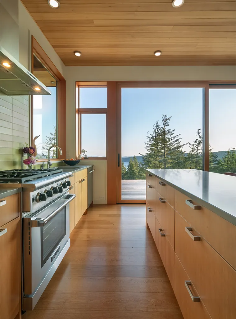 Modern kitchen with wooden floors and cabinets, stainless steel stove, large island, and sliding glass door overlooking trees and hills.