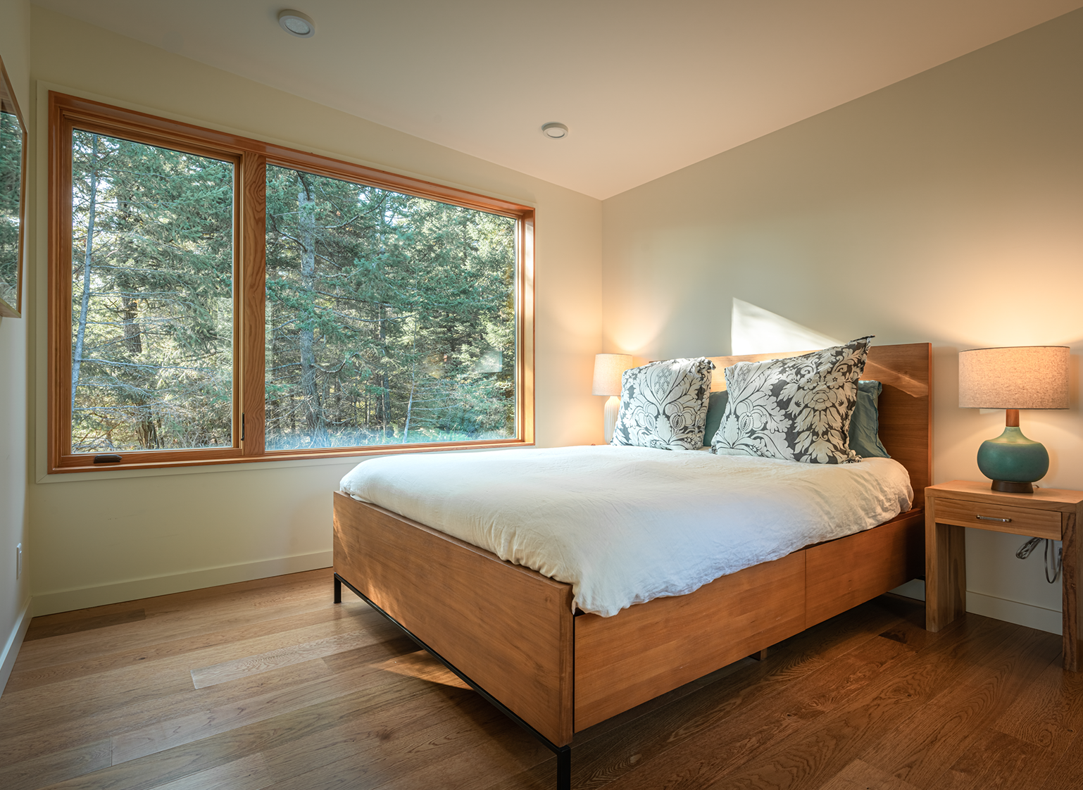 Bedroom with large wooden bed, white bedding, patterned pillows, two bedside tables with lamps, and a large window showing green trees outside.