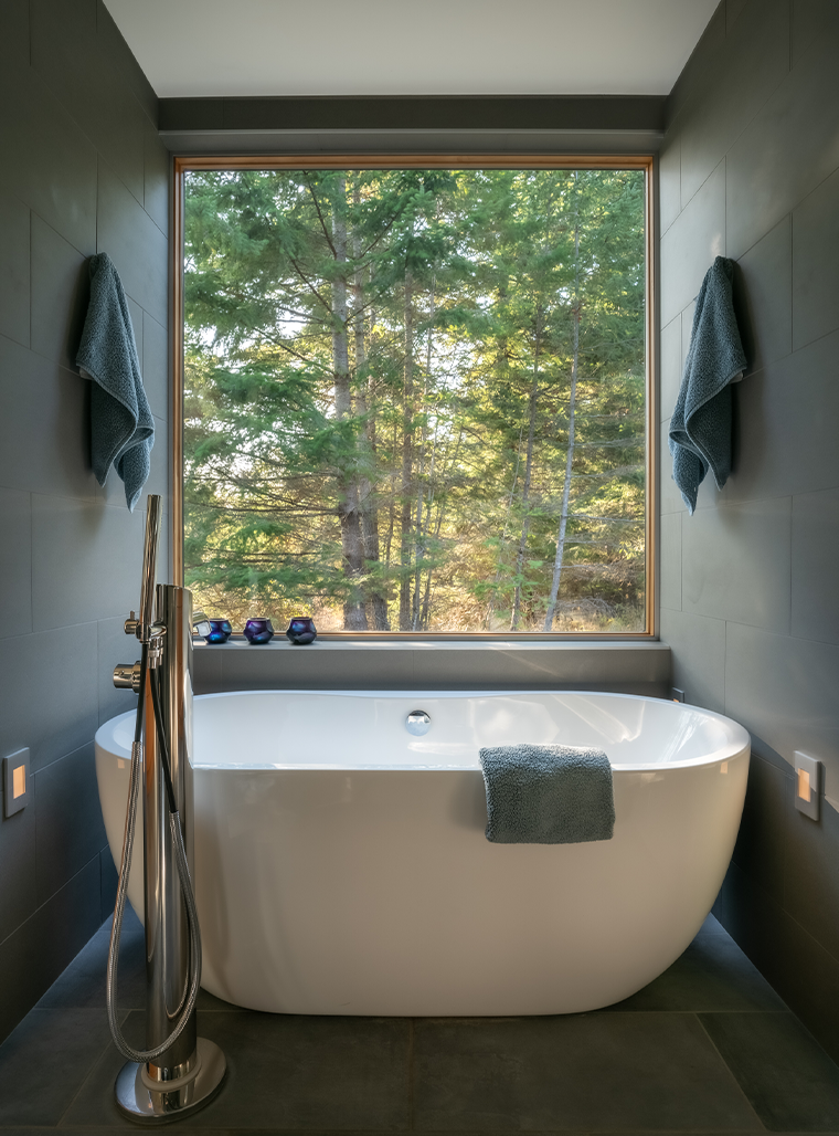 Modern white freestanding bathtub with towels on side and walls, large window showing green forest outside.