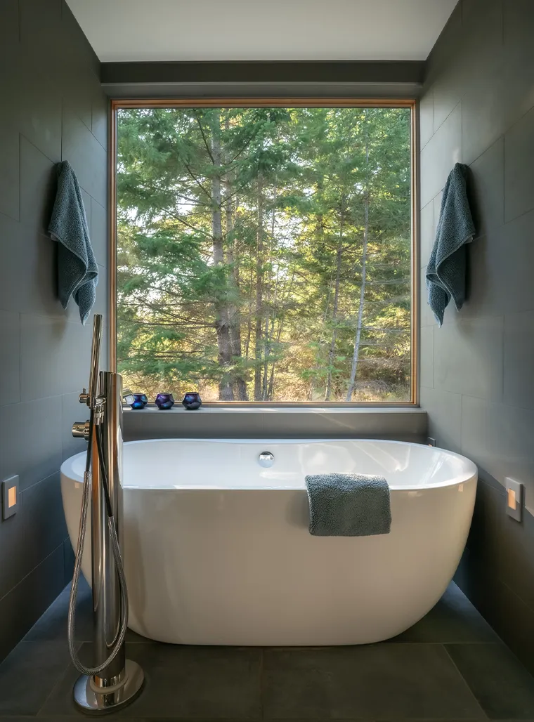 Modern white freestanding bathtub with towels on side and walls, large window showing green forest outside.