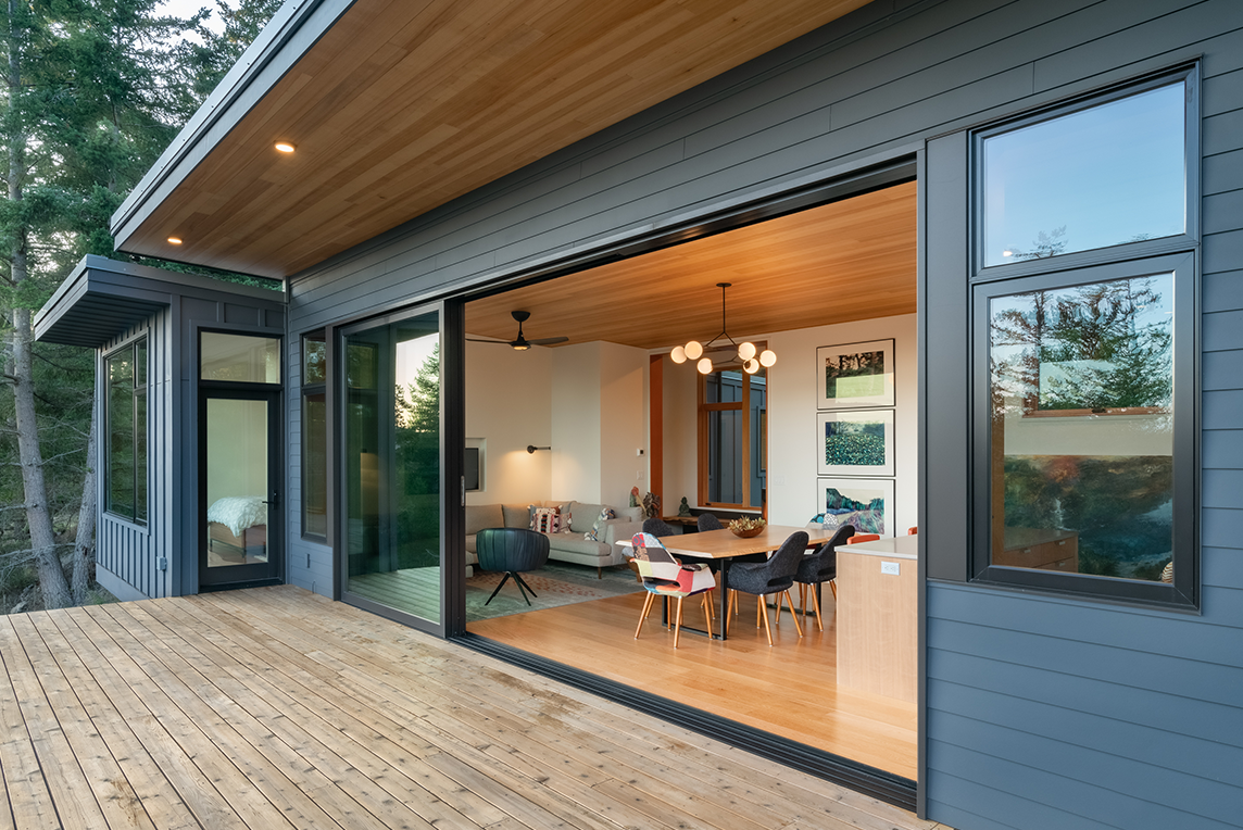 Modern house exterior with large open sliding glass doors revealing a dining area and living room with wood ceiling and floors.