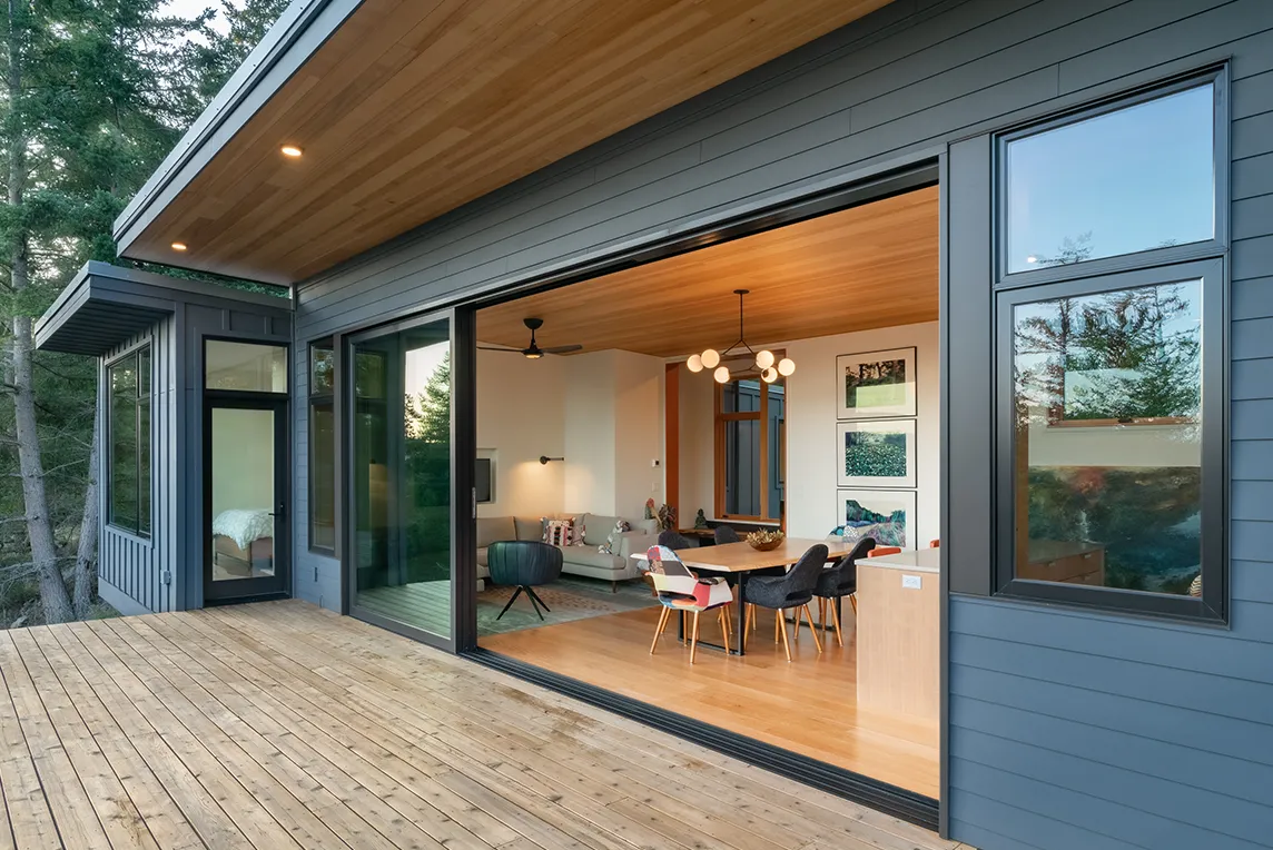 Modern house exterior with large open sliding glass doors revealing a dining area and living room with wood ceiling and floors.