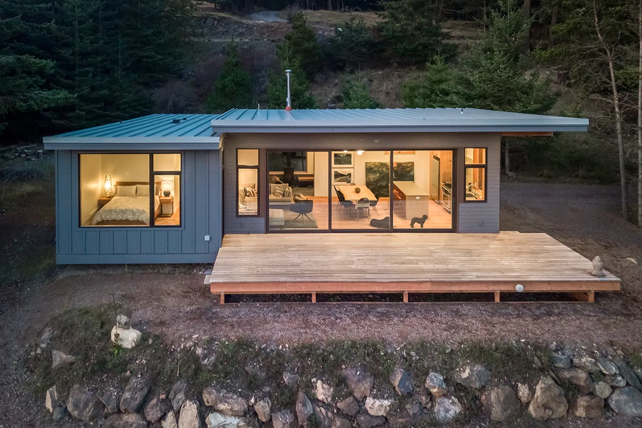 Modern single-story cabin with large windows showing a bedroom, living area, dining table, and kitchen, surrounded by trees and rocky terrain.