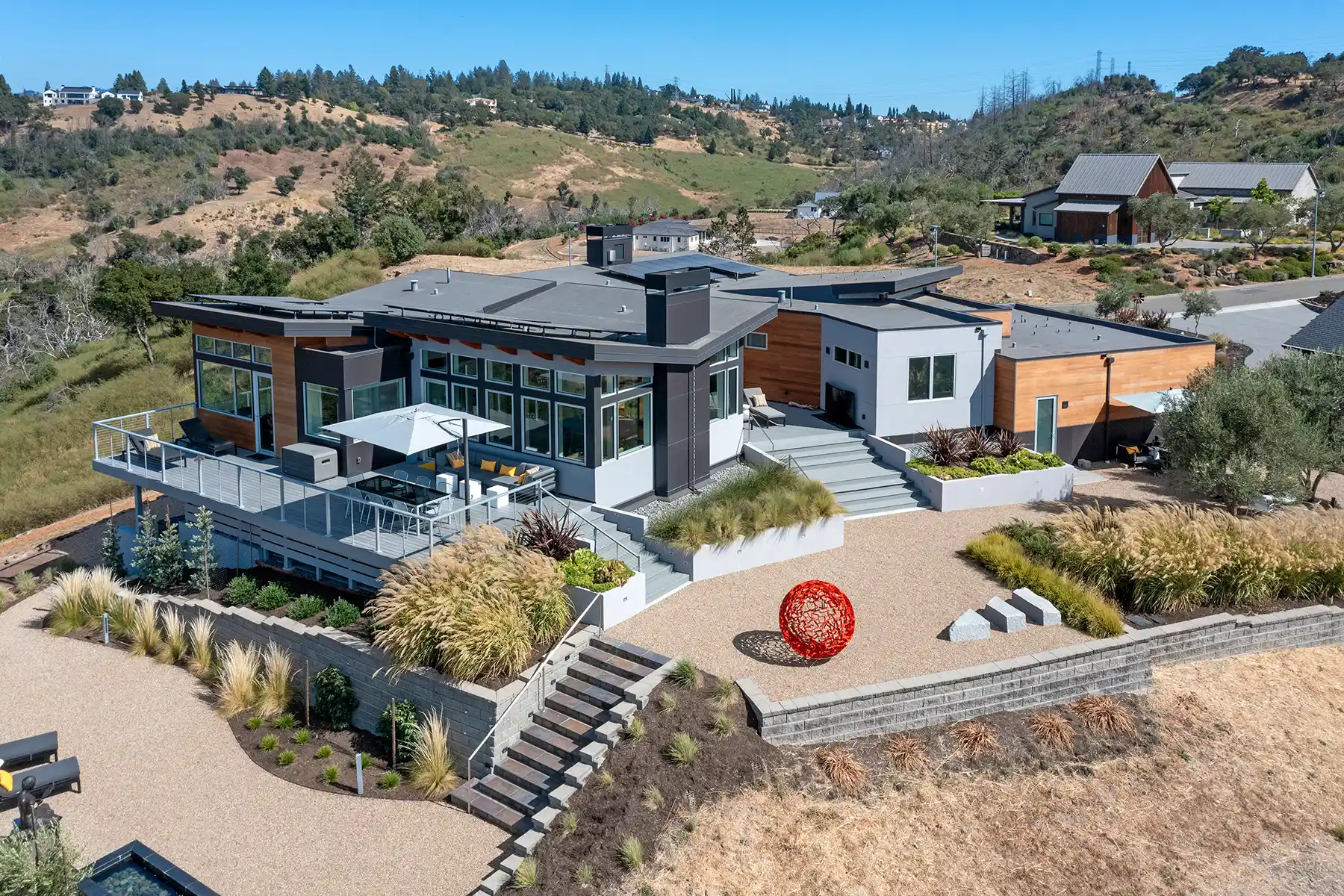 Modern hillside house with large windows, outdoor seating under an umbrella, landscaped garden, and a red spherical sculpture in the gravel courtyard.