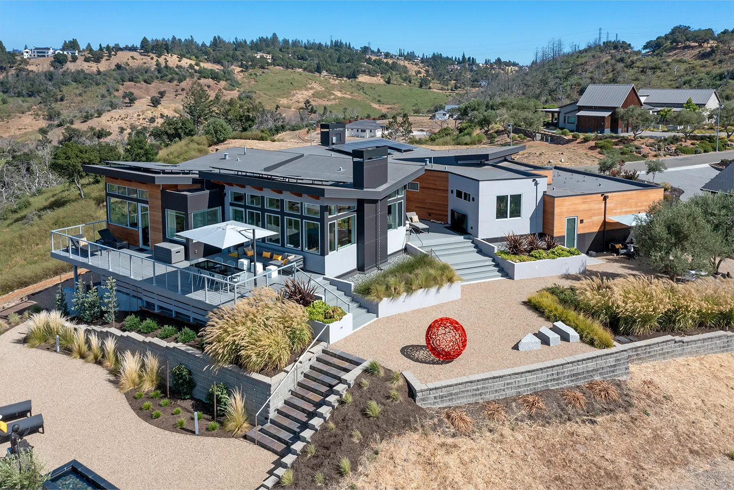 Modern hillside house with large windows, patio furniture under an umbrella, landscaped gravel yard, and a red spherical sculpture.