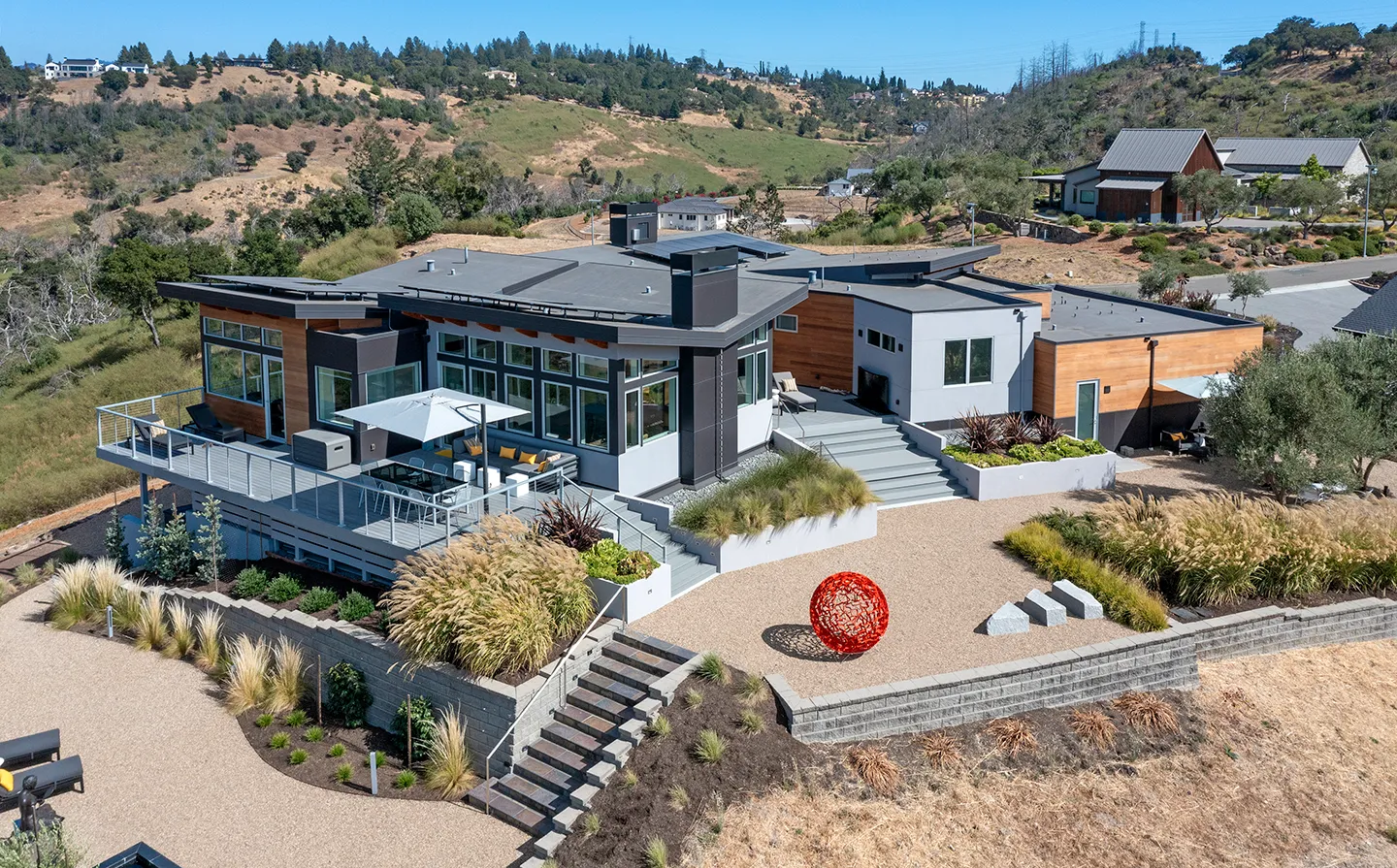 Modern hillside house with large windows, patio furniture under an umbrella, landscaped gravel yard, and a red spherical sculpture.